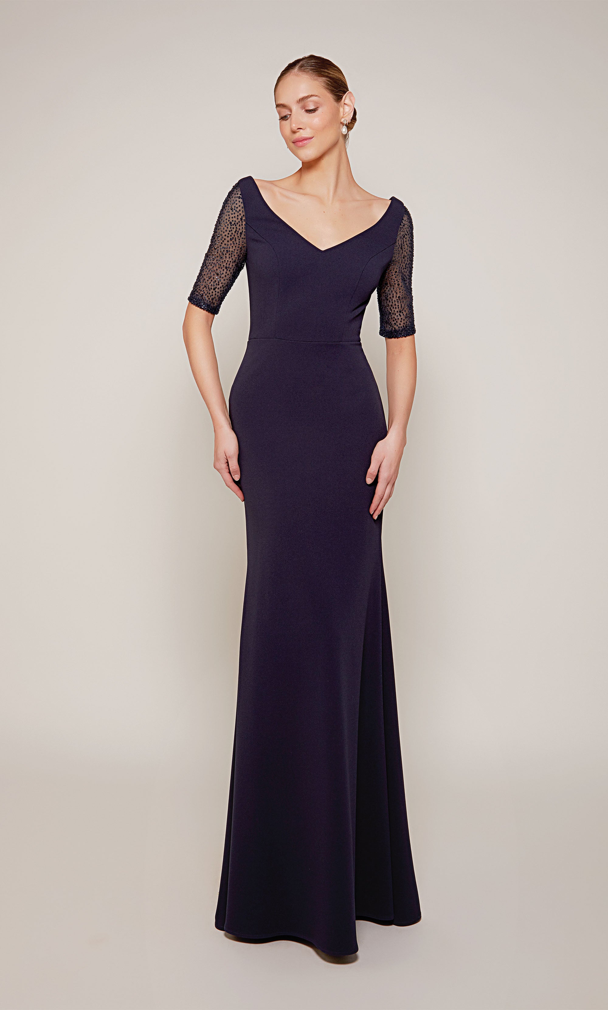 A long navy wedding guest dress with a wide V-neckline, short embellished sleeves, and a fitted skirt that flares slightly from the knee to the bottom of the hemline.