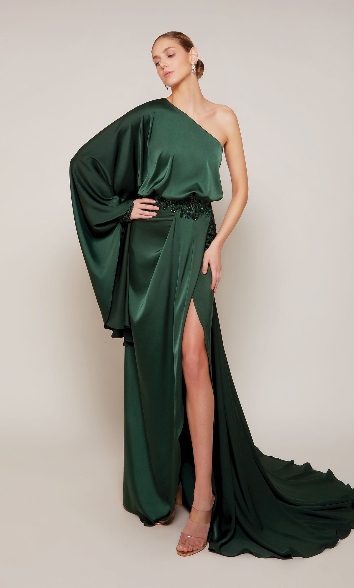 A draped satin evening gown with a one shoulder neckline, beaded waistline, side slit, and train in a rich forest green.