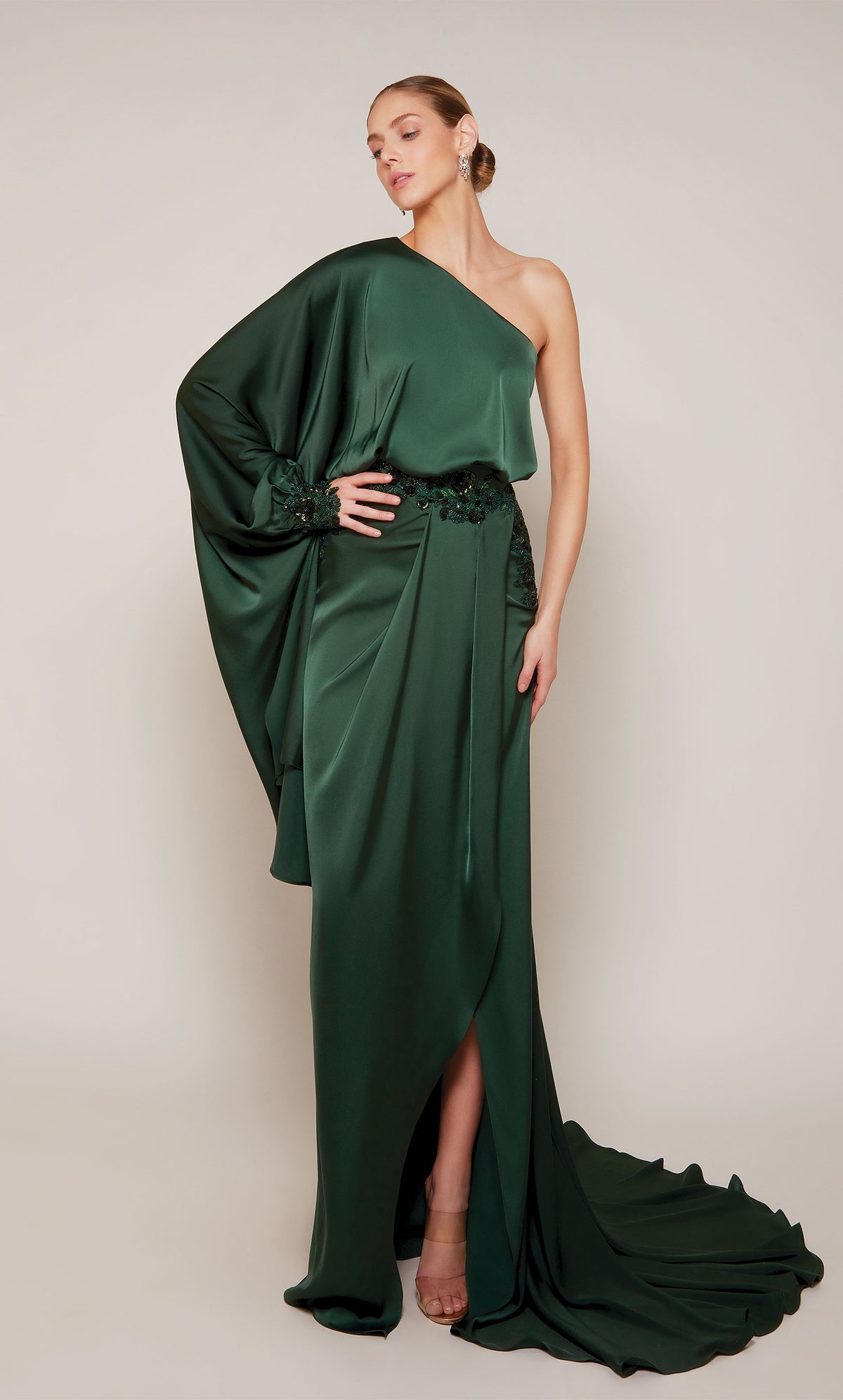 A draped satin evening gown with a one shoulder neckline, beaded waistline, side slit, and train in a rich forest green.