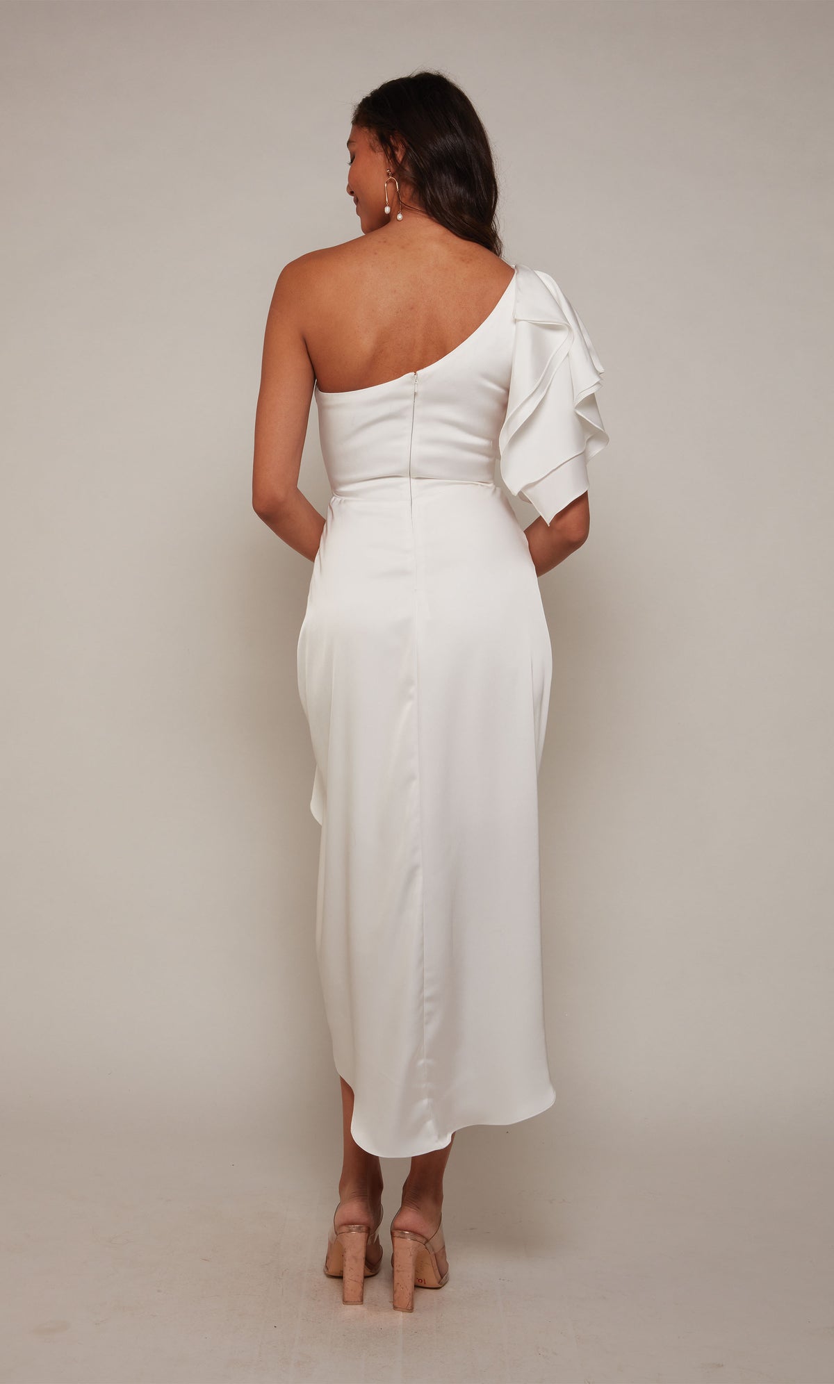 A white one shoulder cocktail dress with an asymmetrical hemline, ruffle shoulder, and a zip-up back closure.