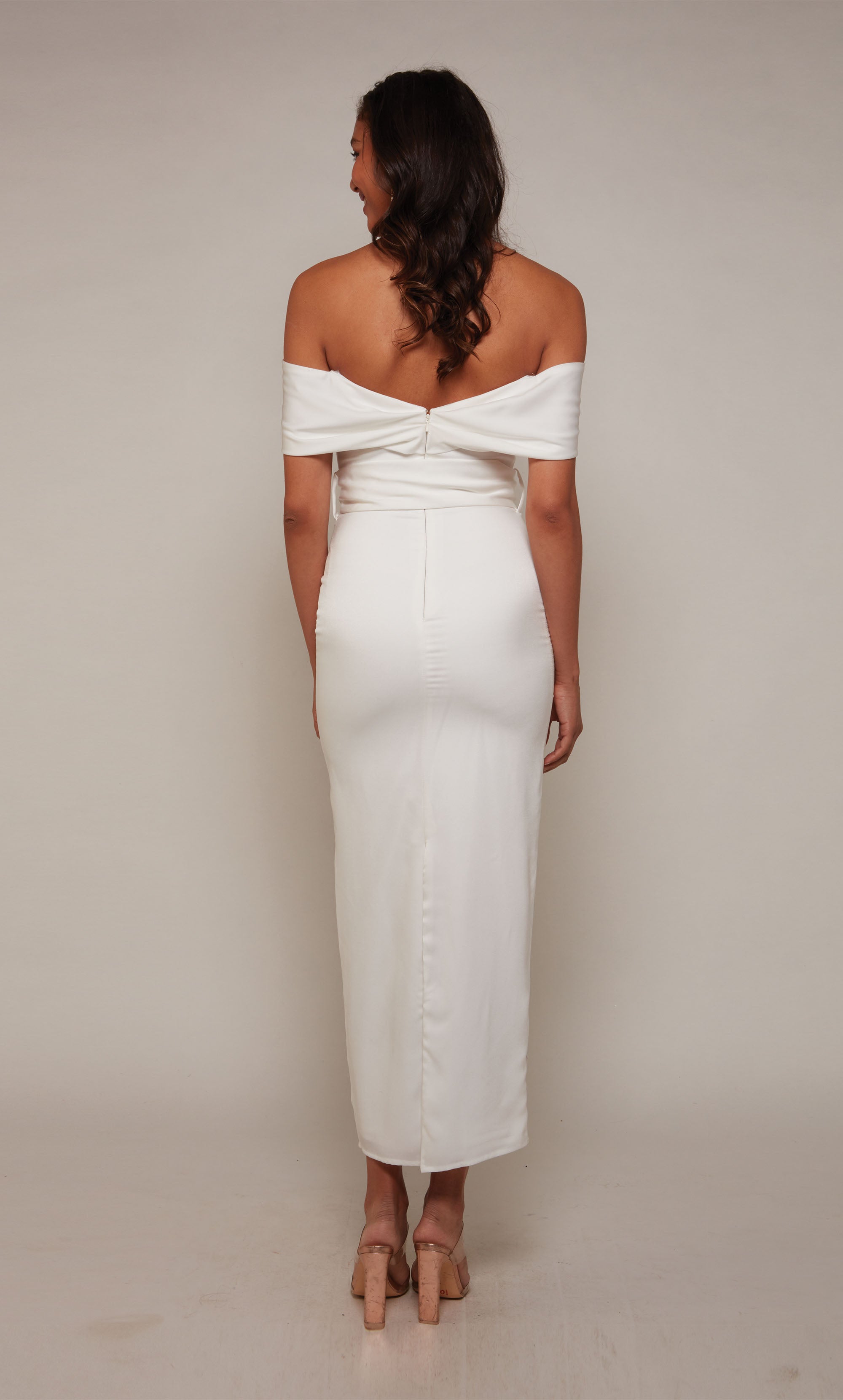 A  white, satin midi dress with an off the shoulder neckline and thick belt at the waistline. The skirt is a fitted pencil skirt with a back slit.