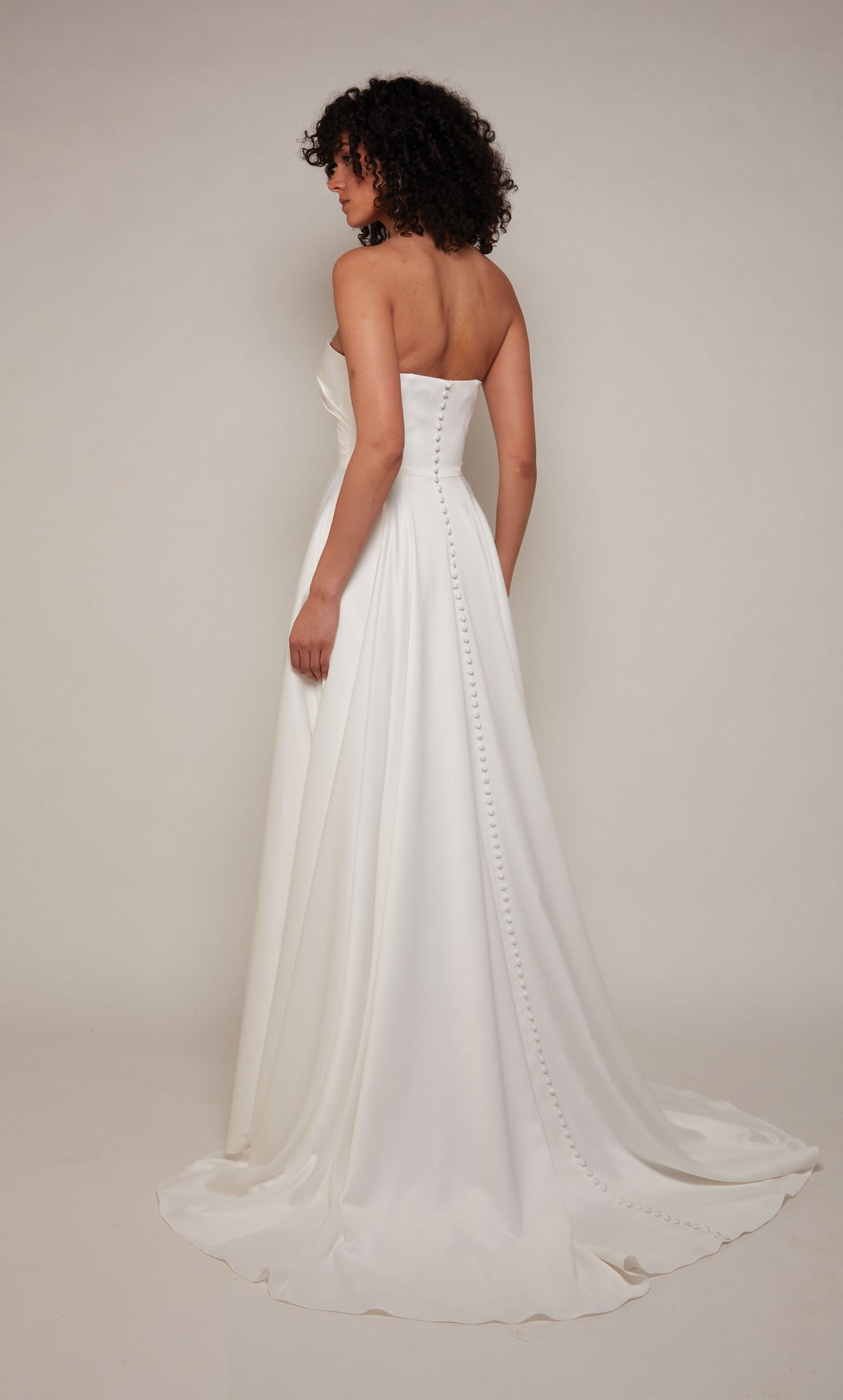 A-line satin bridal gown featuring a strapless, semi-sweetheart neckline, a pleated bodice, and high side slit. Satin covered buttons cascade down the back of the dress from the top to the end of the elegant train.