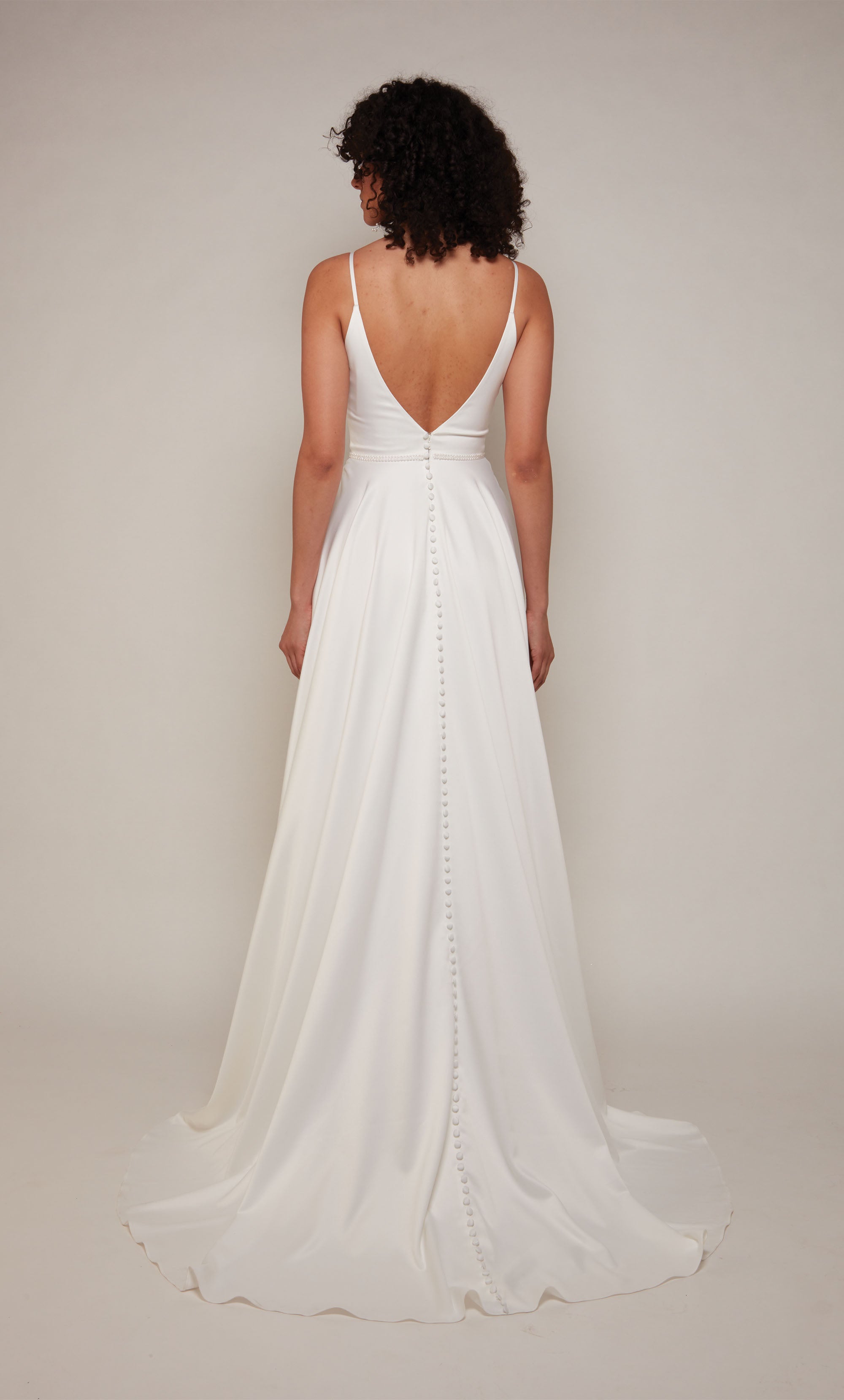 A simple, satin wedding dress with a V-neckline, an A-line skirt, pearl accented waistline, and V-shaped open back with satin covered buttons cascading down the back to the tip of the elegant train.