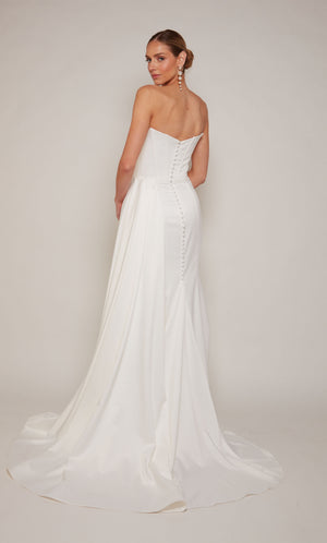 A strapless, corset wedding dress with a ruched waist, an elegant side slit, and detachable side train for a dramatic effect. The back of the dress is adorned with satin covered buttons.
